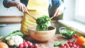 A person mixes a salad in a wooden bowl surrounded by various fresh vegetables