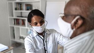 A female Black doctor in a mask listens to a Black male patient's heart using a stethoscope
