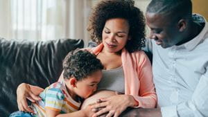 A young boy and young Black man sitting on the couch next to a pregnant Black woman