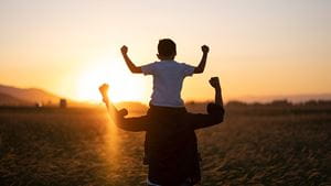 Silhouette of young boy on dad's shoulders flexing muscles as sun sets in front  of them