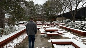 Man in winter jacket walks alone with his head down past snowy picnic tables on his right and pine trees on his left
