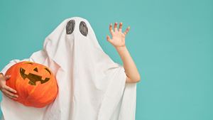 A child in a homemade ghost Halloween costume waves while holding a pumpkin in front of a teal background