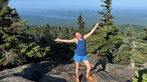 Jolene with her arms out on Jenkins Mountain, surrounded by pine trees and a view of an Adirondack lake