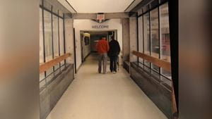 A man using a walker walks alongside another man down a hospital hallway