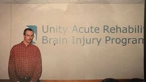 A white man with a mustache wearing a red and white checkered shirt standing in front of a white wall that says Unity Acute Rehabilitation Brain Injury Program in blue lettering