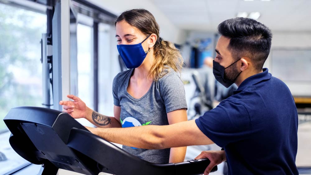 A man wearing a mask helps a woman wearing a mask who is walking on a treadmill