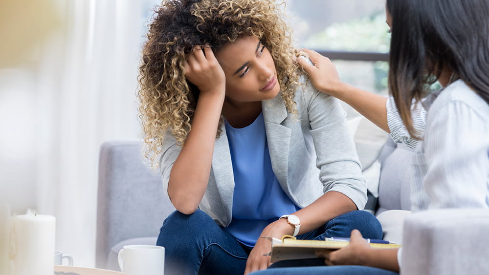 A female therapist places a hand on the shoulder of a young Black woman to comfort her